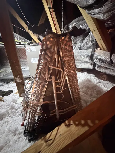 New copper evaporator coil standing upright in an attic between wooden rafters, ready to be installed with the air handler